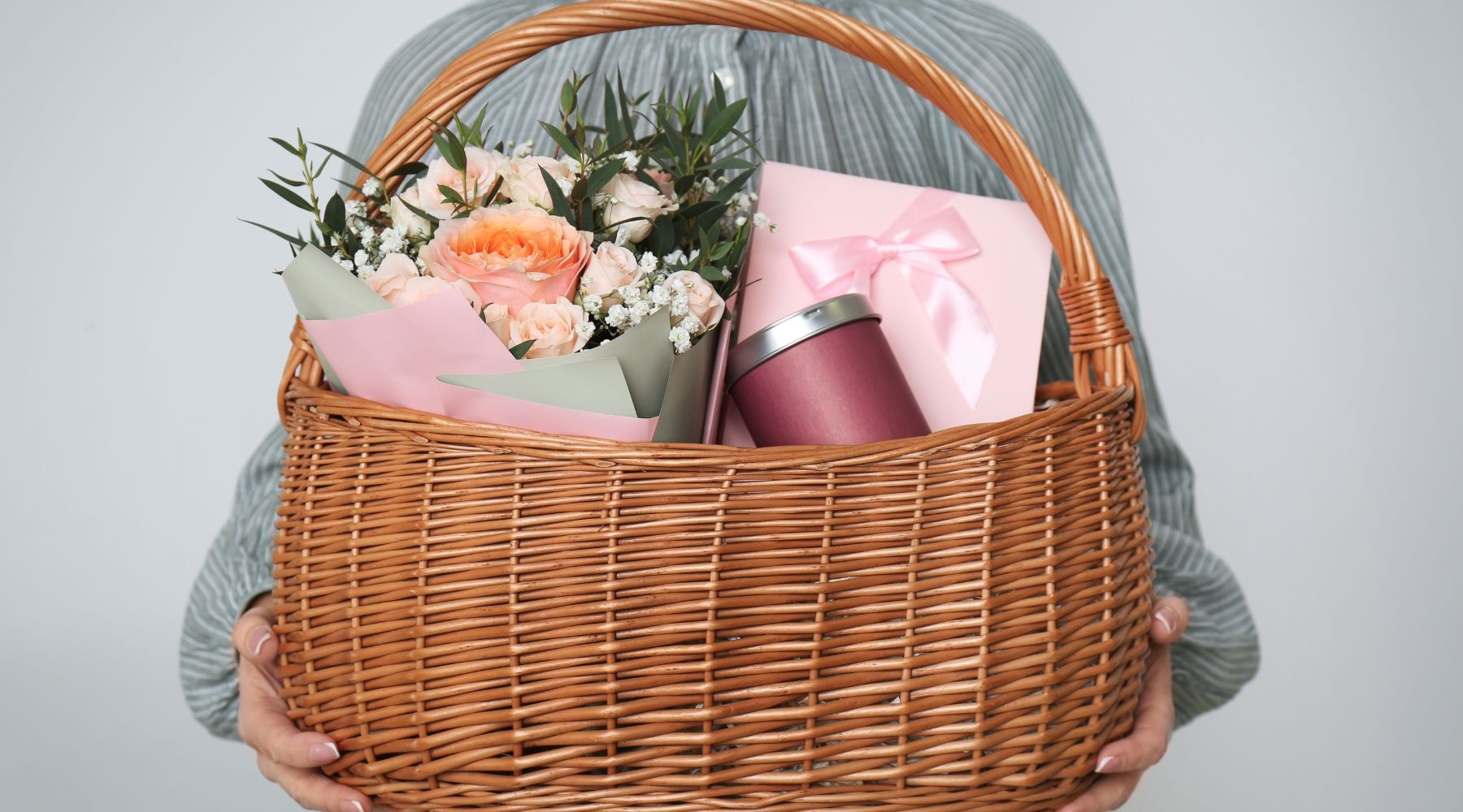 Wicker basket held by person, containing flowers, cards, and colored tins.