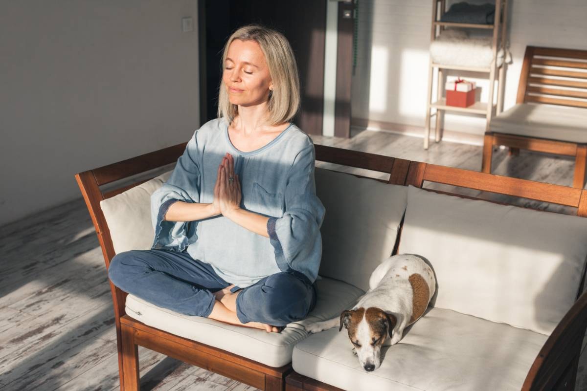 A woman meditates on a couch with her dog resting beside her, highlighting Tips for Beginners in Meditation for Dogs