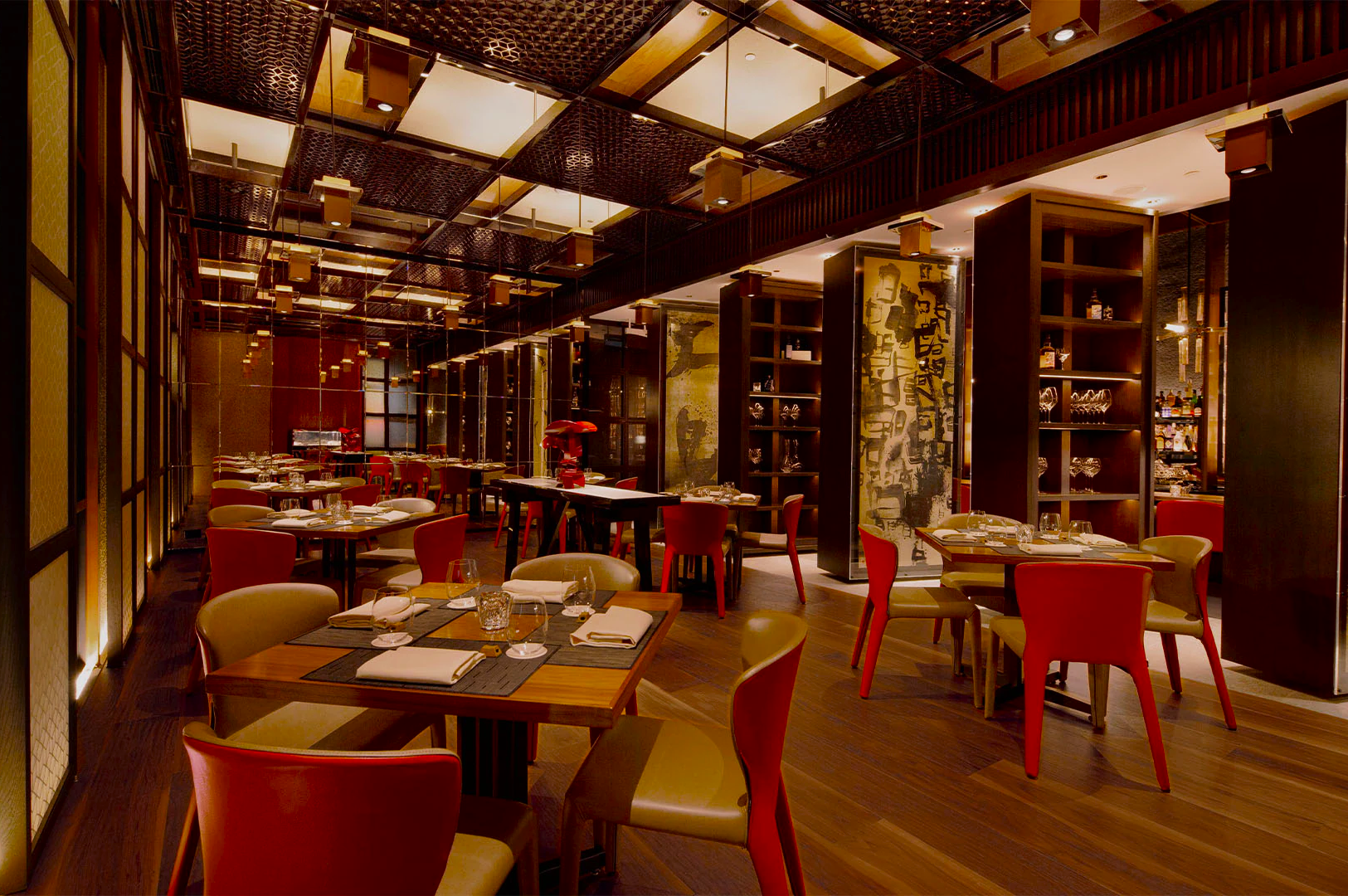 Elegant restaurant interior with wooden floors, red and beige chairs, and warmly lit tables set with glassware and napkins. Cozy and intimate ambiance.