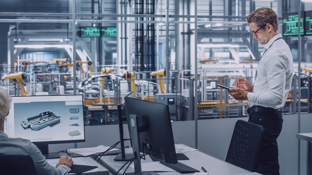 Modern automotive production facility – two employees in the foreground, one using a computer with a visible 3D model, the other interacting with a tablet; robotic arms on the automated assembly line in the background – illustrating digitalization enabled by PTC MPMLink and Windchill.