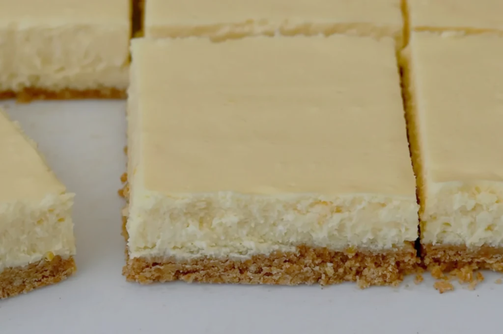Close-up of creamy cheesecake bars with a smooth top and a crumbly brown crust, cut into squares, sitting on a white surface.