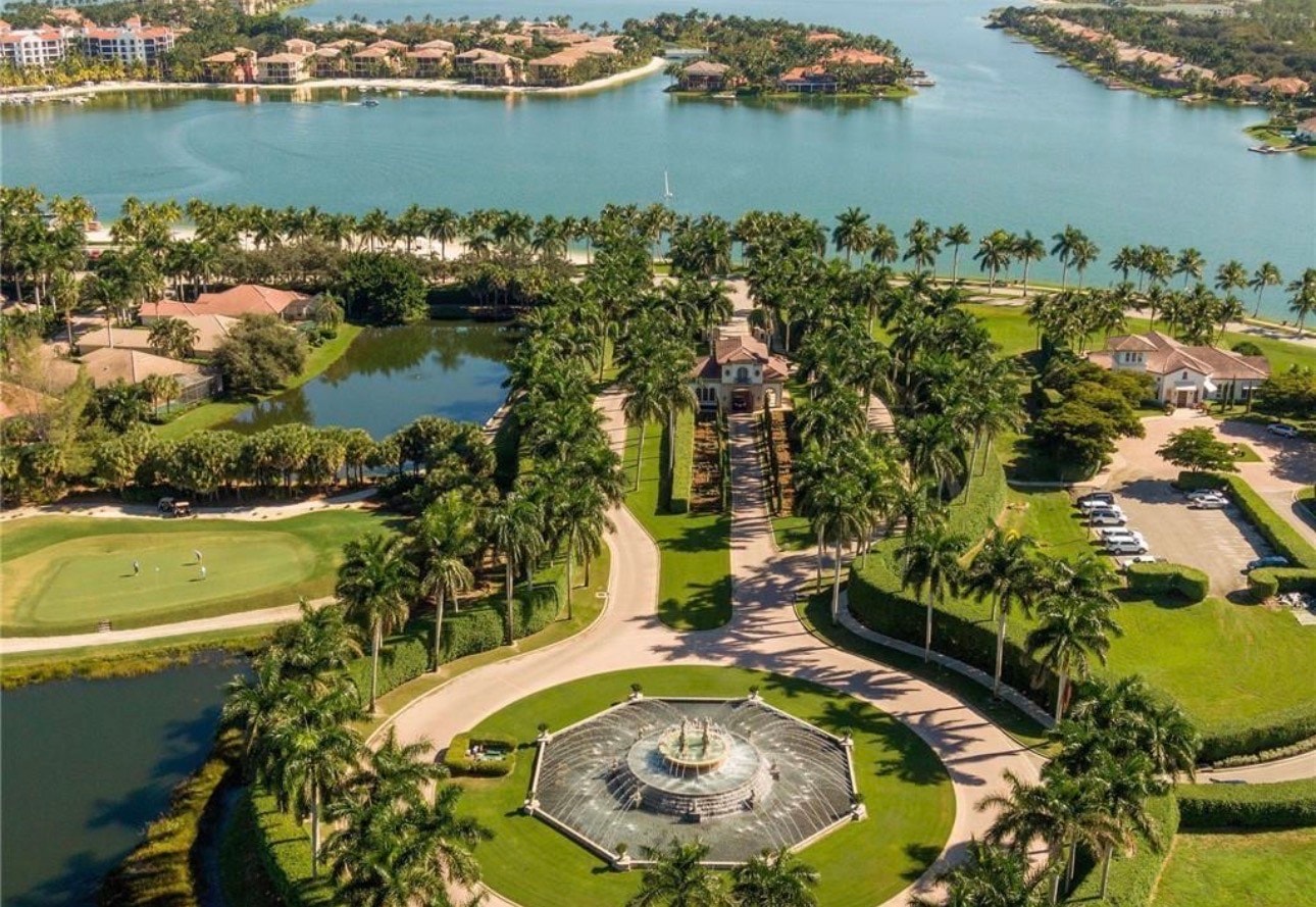 The image depicts a serene lakefront dining area with outdoor seating, offering a breathtaking view of the lake at sunset. This tranquil setting is part of the luxurious Miromar Lakes Beach and Golf Club, where residents can enjoy fine dining and picturesque moments over the contiguous aquamarine freshwater lakes.