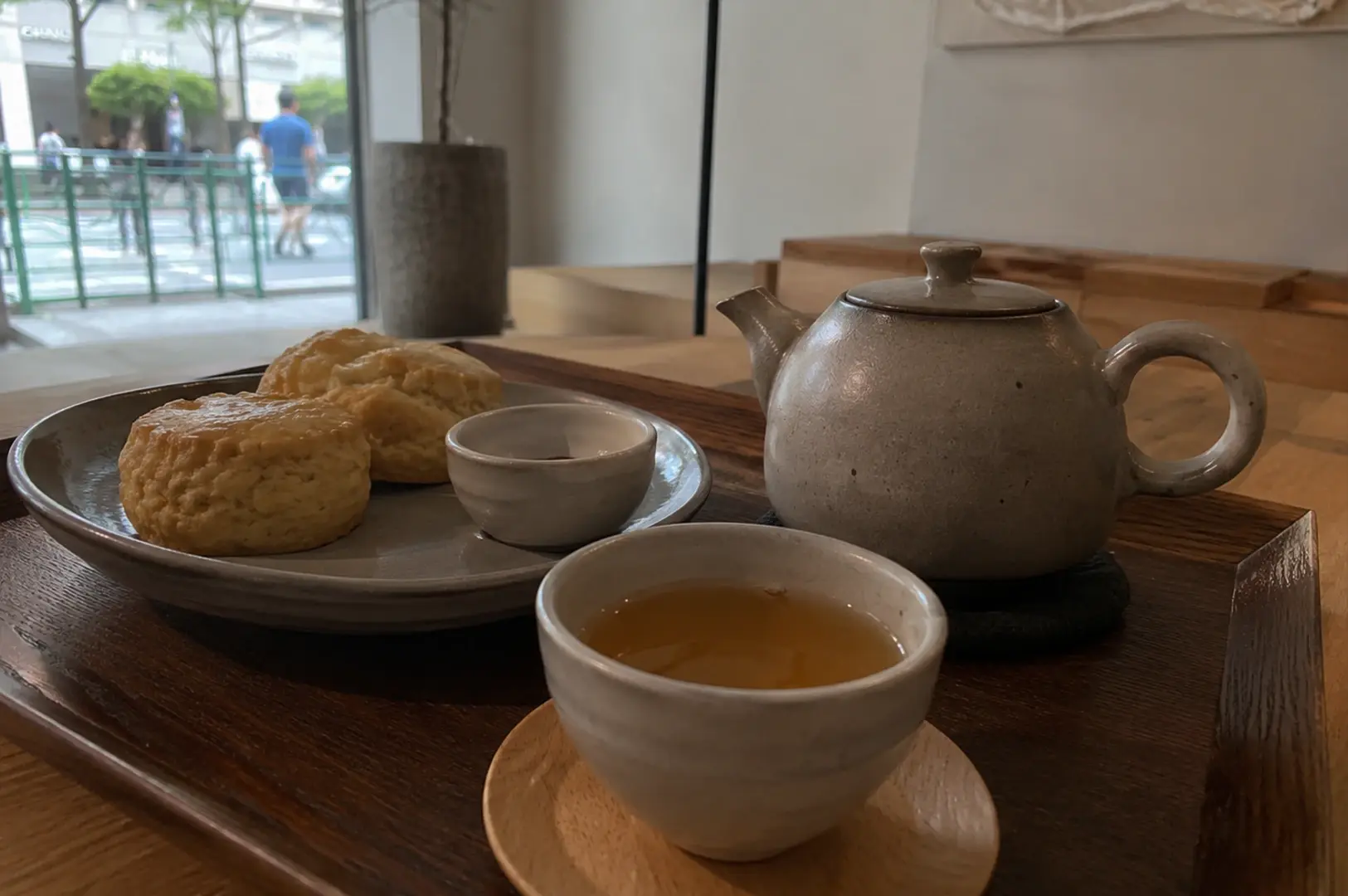 An eye-level close-up shows a tea and snack serving on a dark wooden table inside a modern café. In the foreground, a smooth, light-grey ceramic cup filled with amber-colored tea rests on a round wooden saucer. Behind it to the right sits a large, speckled grey ceramic teapot with a prominent handle and spout, resting on a dark coaster. To the left, a grey ceramic plate holds two golden-brown scones alongside a small dipping bowl. The background is softly blurred, showing wooden bench seating and a large window looking out onto a city street where pedestrians walk past a green railing.