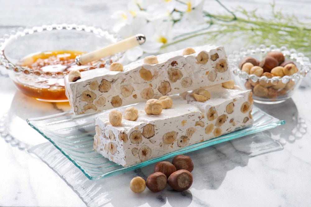 In the image, the production of torrone del torronificio Trapani is captured, showcasing the traditional process of making this Sicilian sweet. Ingredients such as almonds, pistachios, and honey are being mixed in a large pot, while the warm, sticky mixture is prepared to be poured onto a sheet of parchment paper, highlighting the craftsmanship behind this beloved dolce.