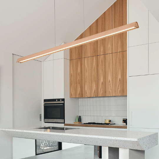 Modern kitchen interior with a long, slim wooden pendant light hanging above a white stone island, featuring wood-paneled cabinets and built-in oven.