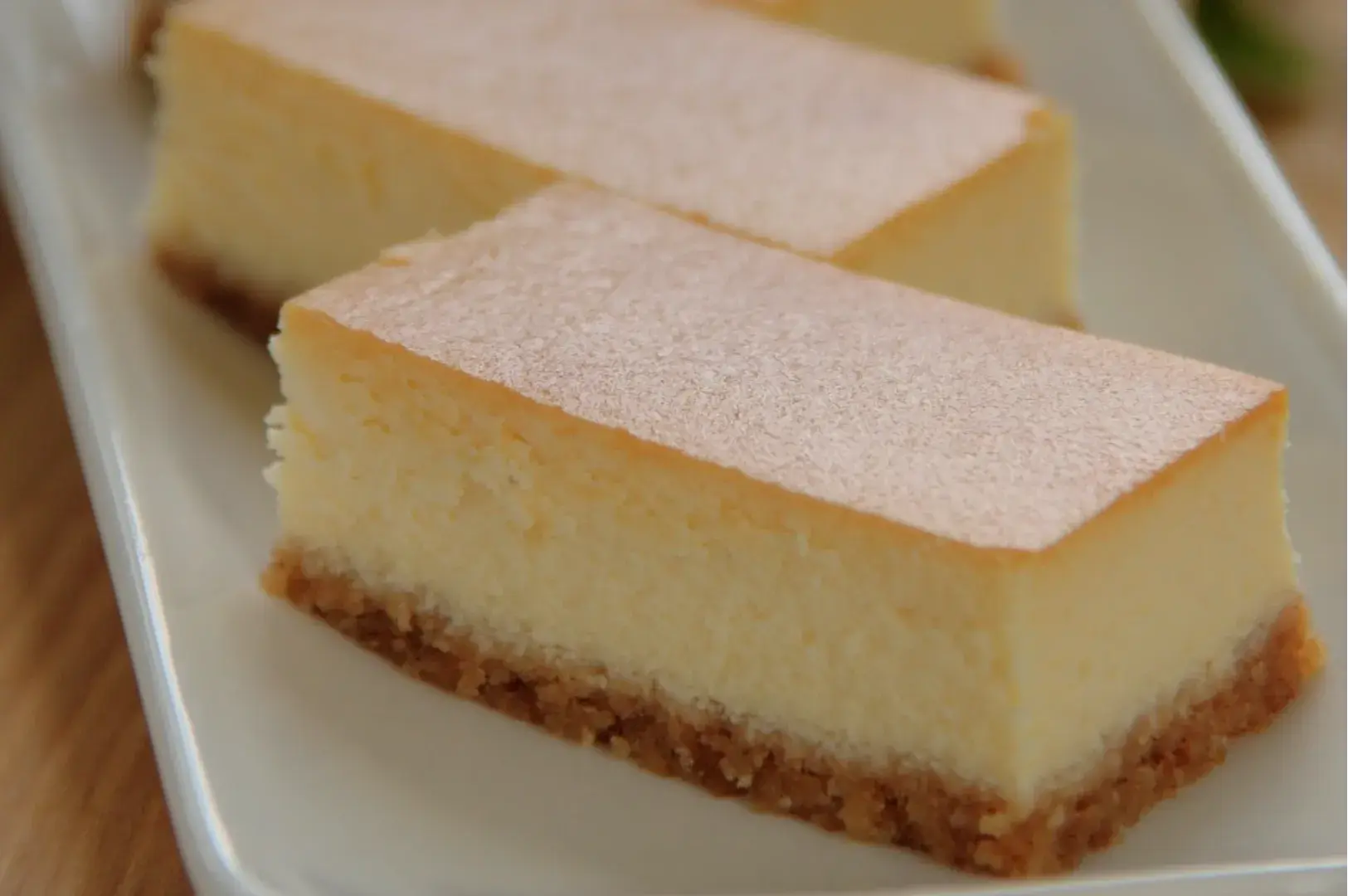 A close-up of slices of cheesecake on a white plate, featuring a smooth creamy layer on top and a crumbly crust at the bottom.