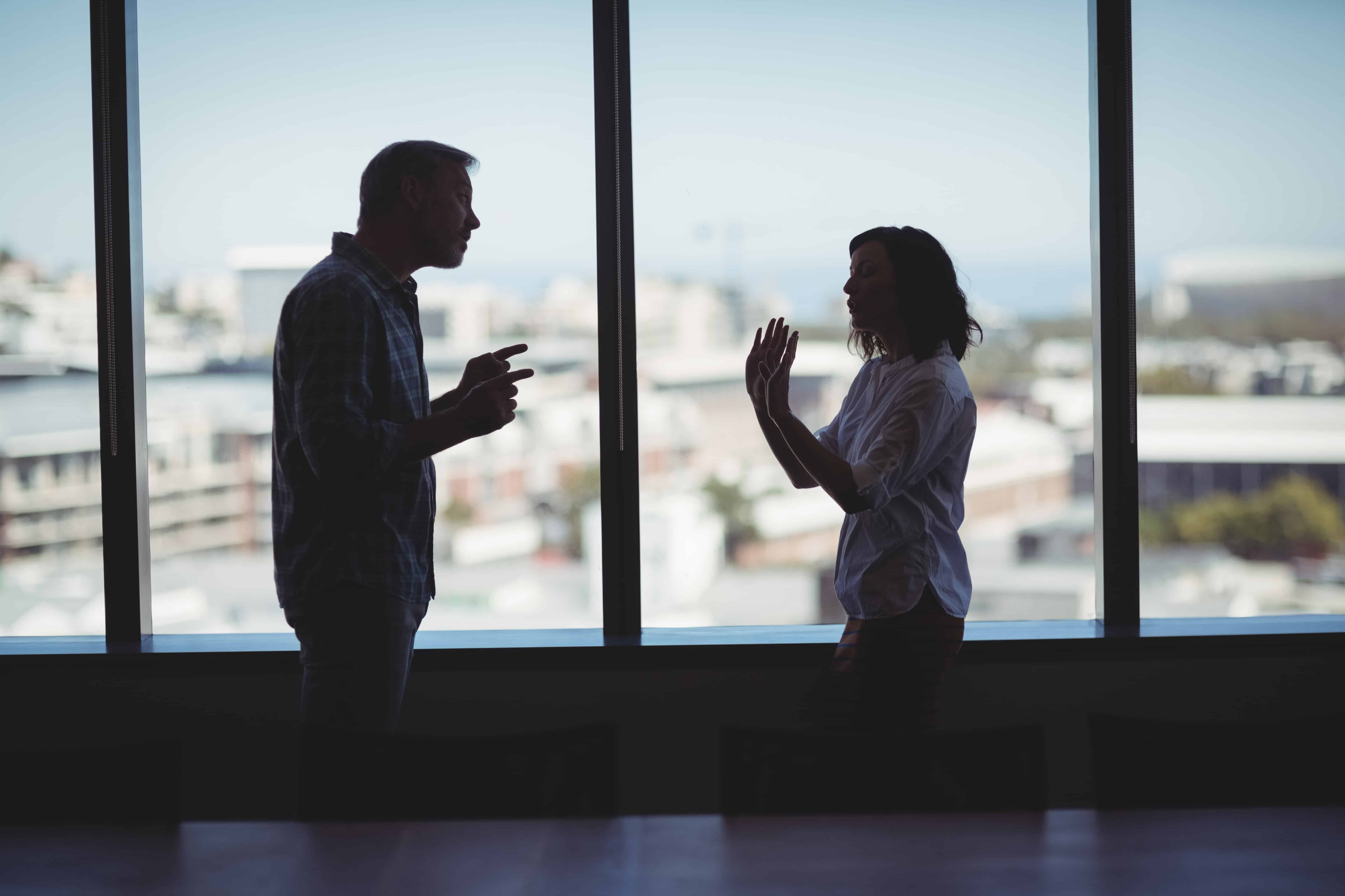 A couple in an office yelling at each other. The man is pointing his fingers to the woman, while she has both her hands up indicating that she doesn't want to argue.
