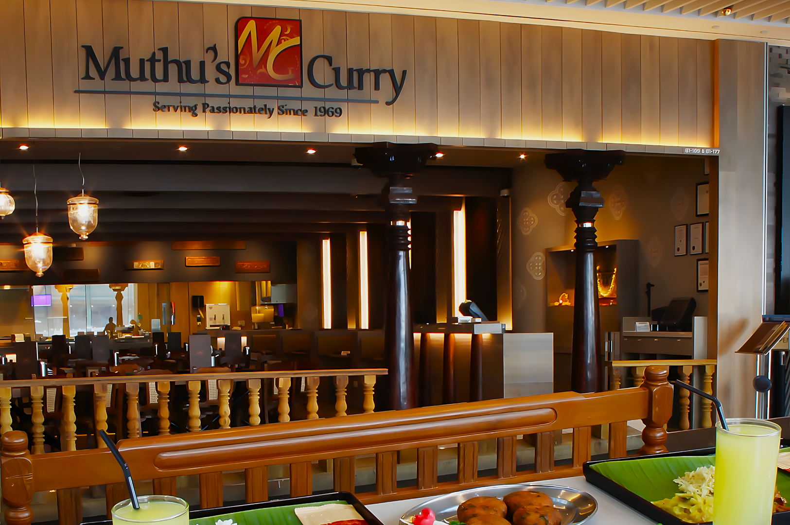 Restaurant interior with wooden decor, warm lighting, and a sign reading "Muthu's Curry." Two trays with food and drinks are visible in the foreground.
