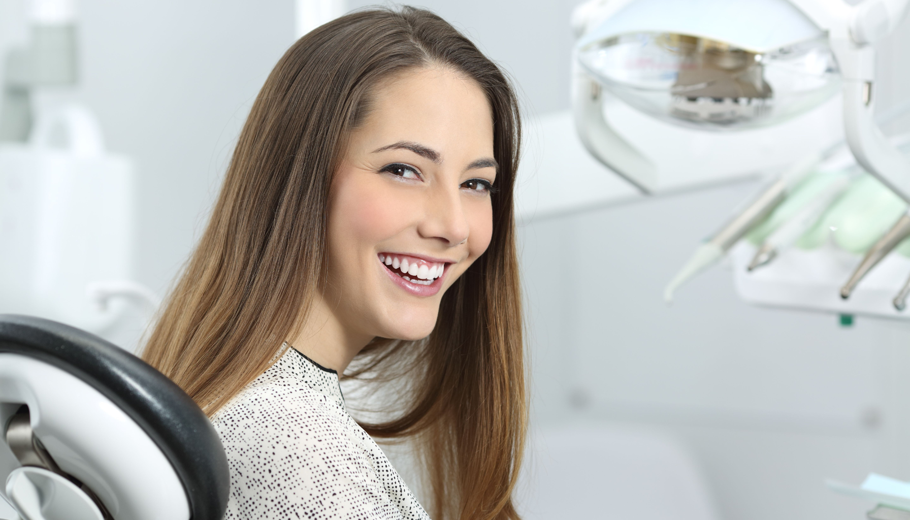 Ung kvinne med langt hår smiler i en moderne tannklinikk, sittende i tannlegestol før behandling.