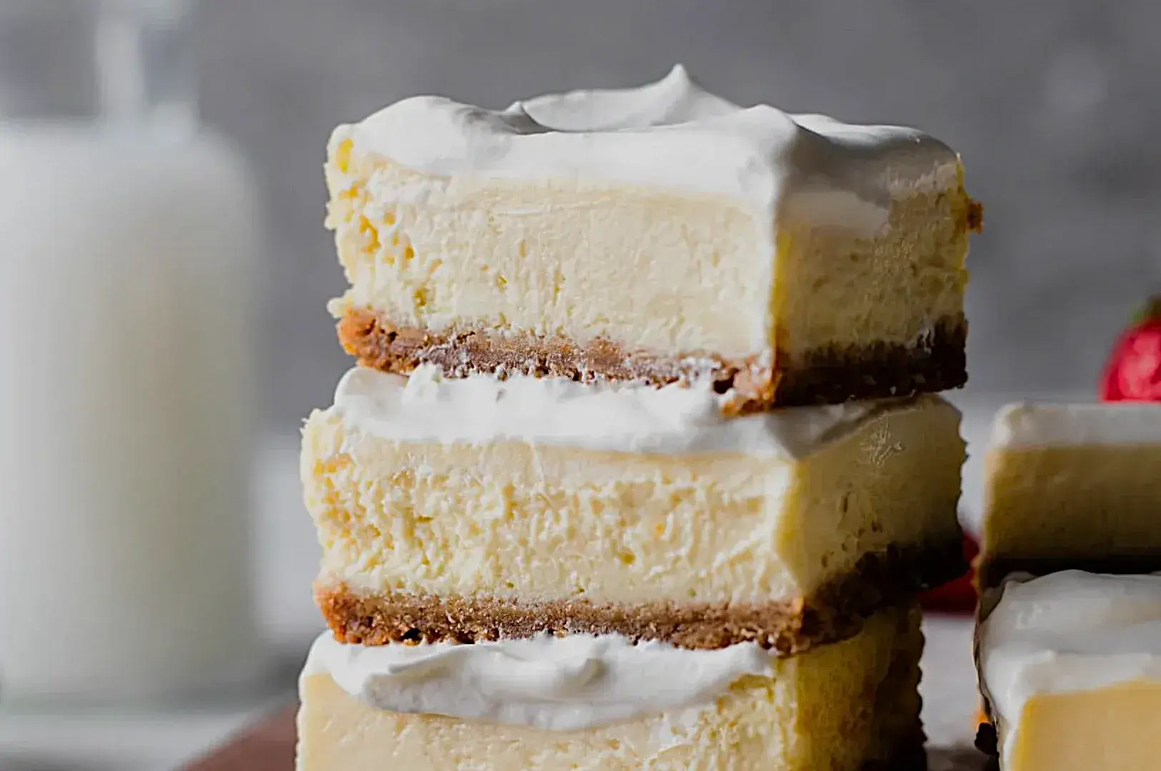 Three layers of creamy cheesecake bars topped with whipped cream, stacked on a wooden board, with a glass of milk in the background.