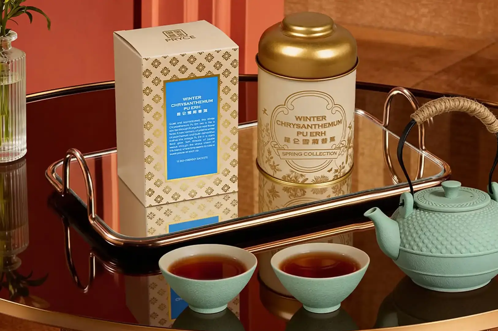 A copper-rimmed mirrored tray displays a white-and-gold patterned tea box alongside a matching white canister labeled "WINTER CHRYSANTHEMUM PU ERH." In front of the tray, two small mint-green ceramic teacups filled with reddish-brown tea rest beside a light mint-green textured teapot on a reflective tabletop.