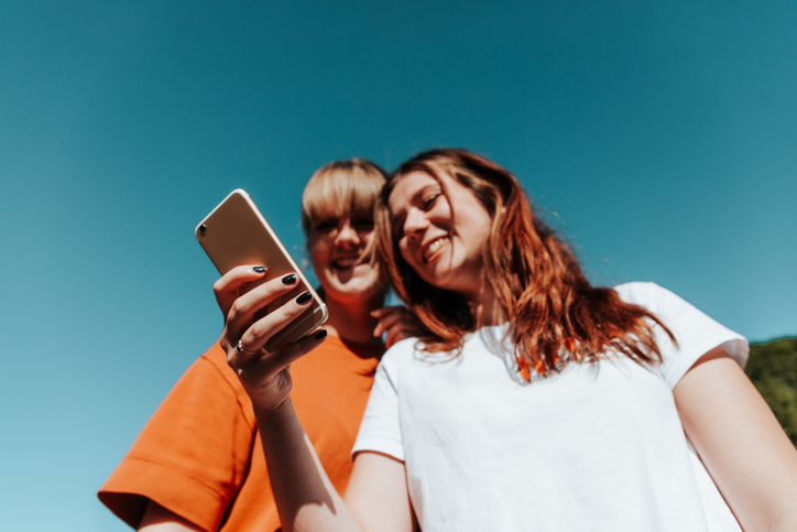 Two young women, one in an orange tee shirt, smiling and looking at a cell phone.