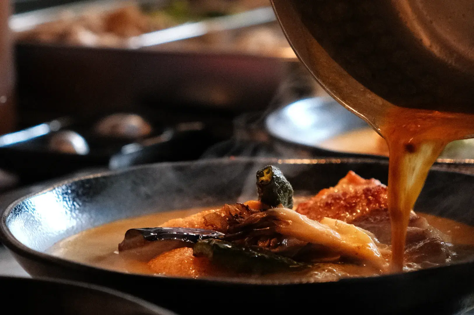 A close-up of a bowl of soup curry with broth being poured over various ingredients, including greens and chicken.