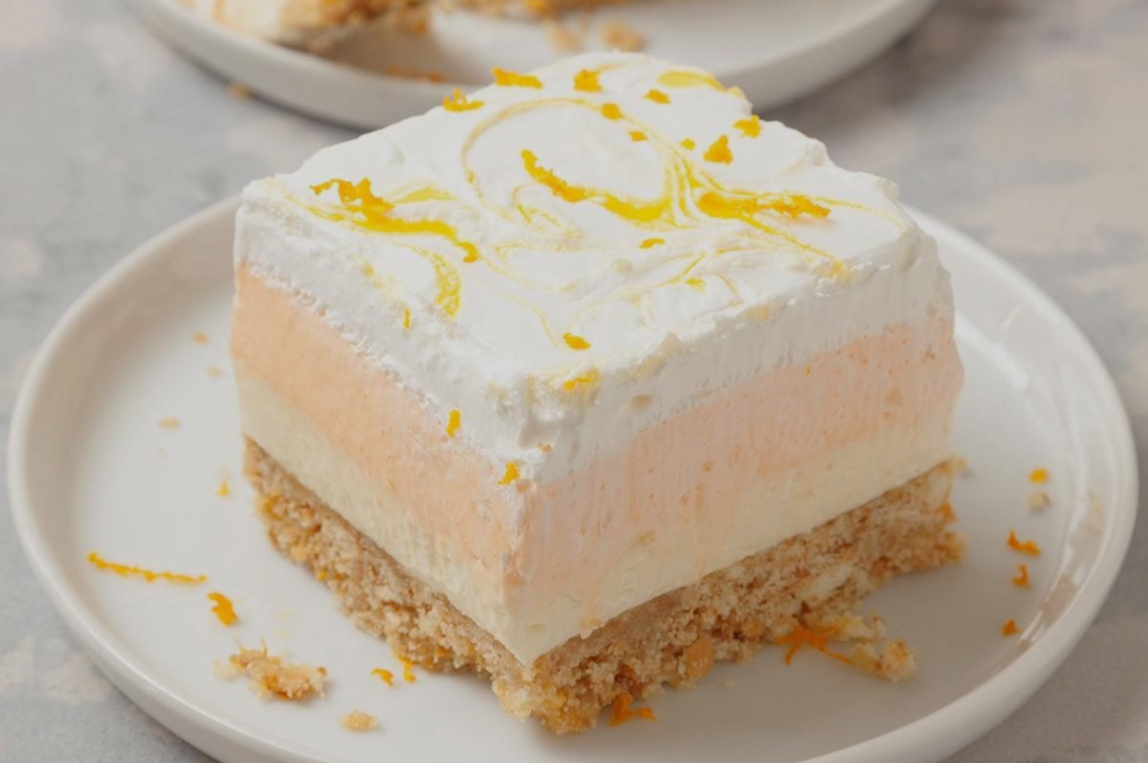 Slice of citrus dessert with a crumbly base, layered with creamy white and orange filling, topped with whipped cream and orange zest on a white plate.
