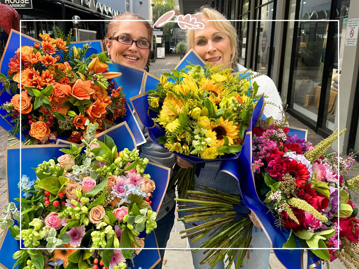 Two women standing outside, each holding multiple vibrant floral bouquets wrapped in blue paper, smiling at the camera - Fabulous Flowers and Gifts