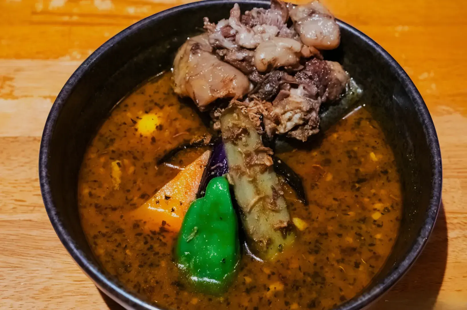 A bowl of savory soup with meat and various vegetables, including green and orange pieces, in a dark broth.