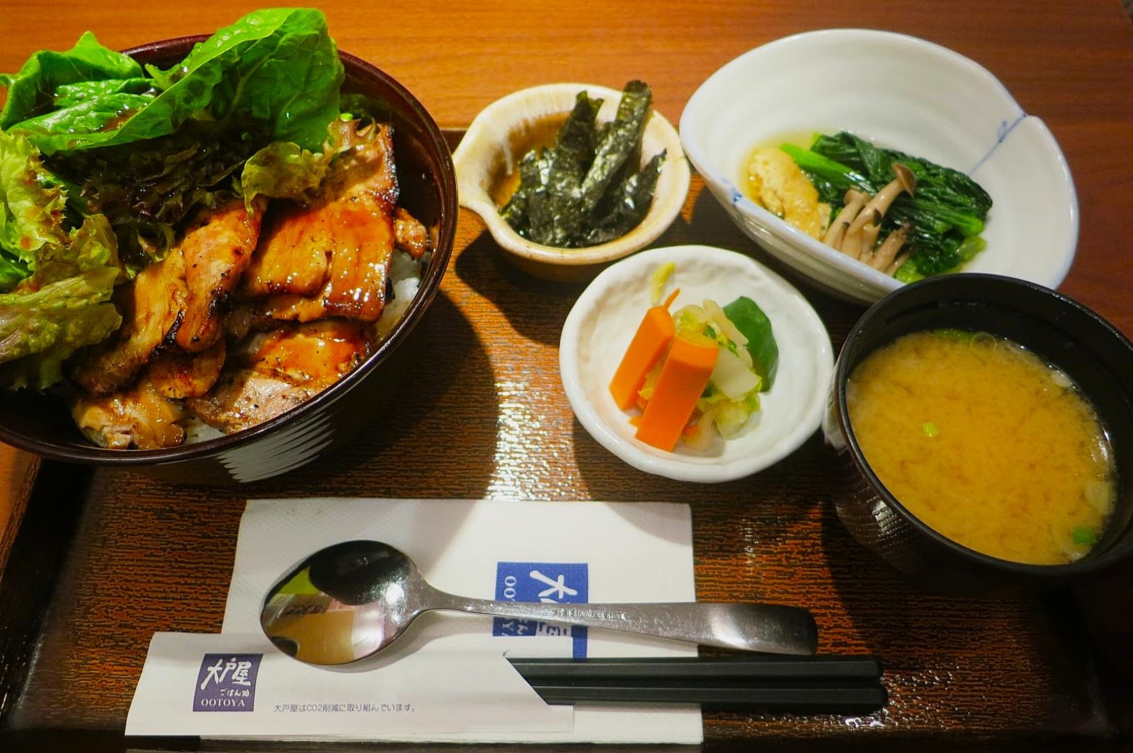 A Japanese meal set with grilled meat and lettuce on rice, seaweed, vegetables, and miso soup. Utensils are placed on a wooden table.