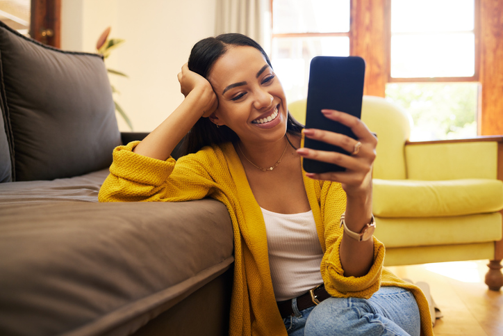 Young woman smiling and sitting on the floor reading something on her cell phone.  