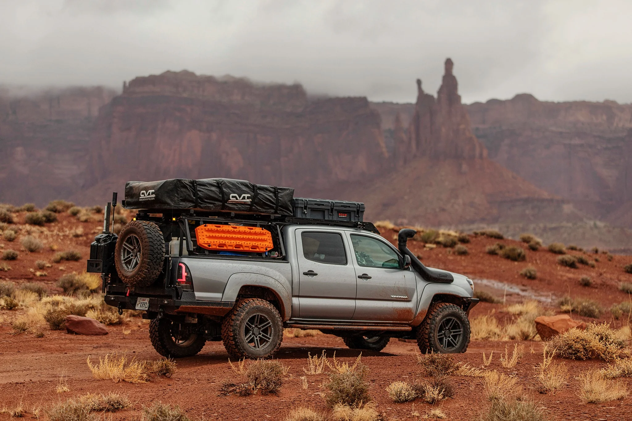 Grey Tacoma with off-road tires, overlanding gear, fuel cans, and tent rack in Utah desert.