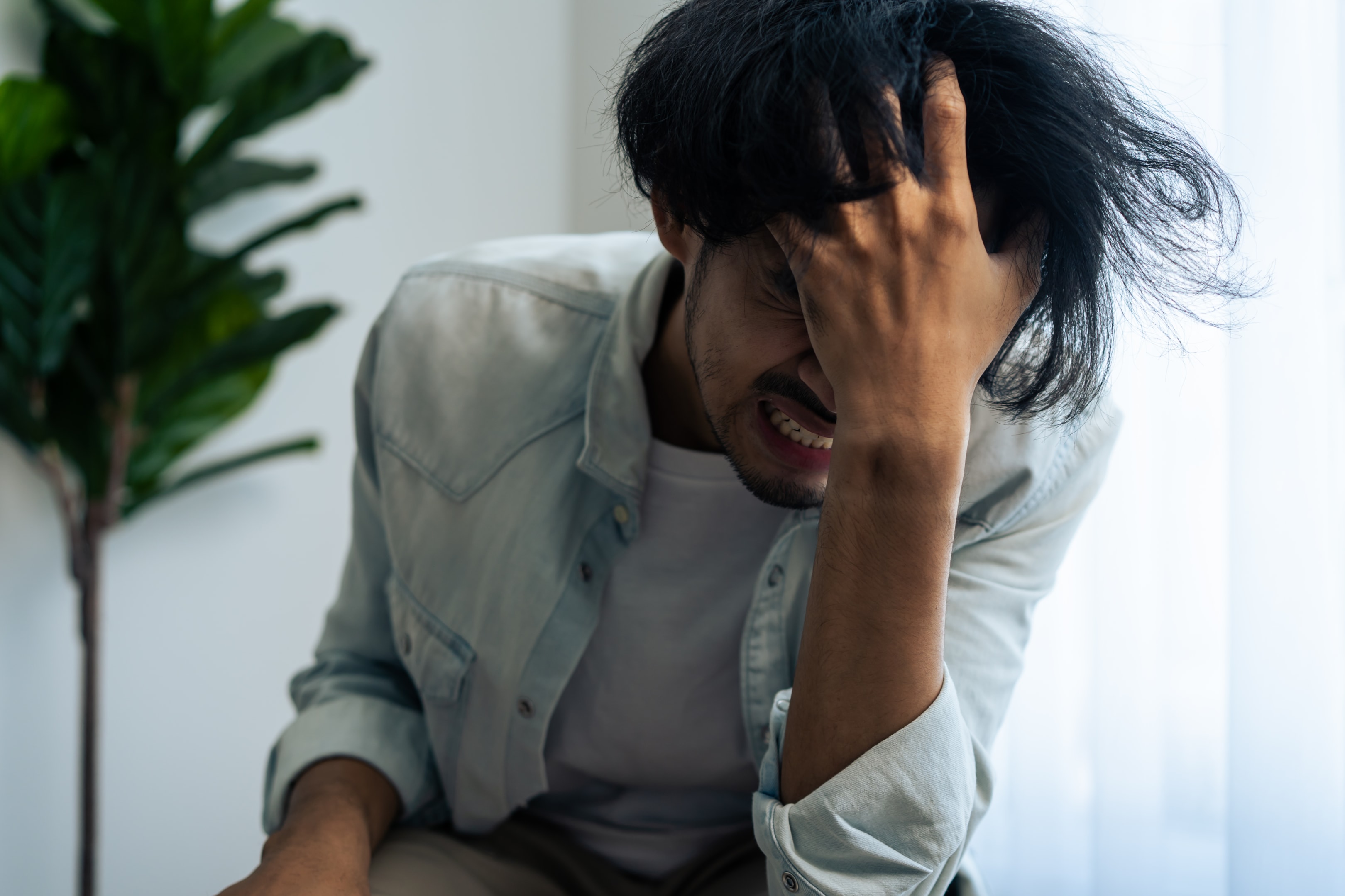A man clutching his forehead in deep distress while sitting near a window, capturing the overwhelming physical and mental intensity that requires the immediate use of Managing Panic Attacks in The Woodlands