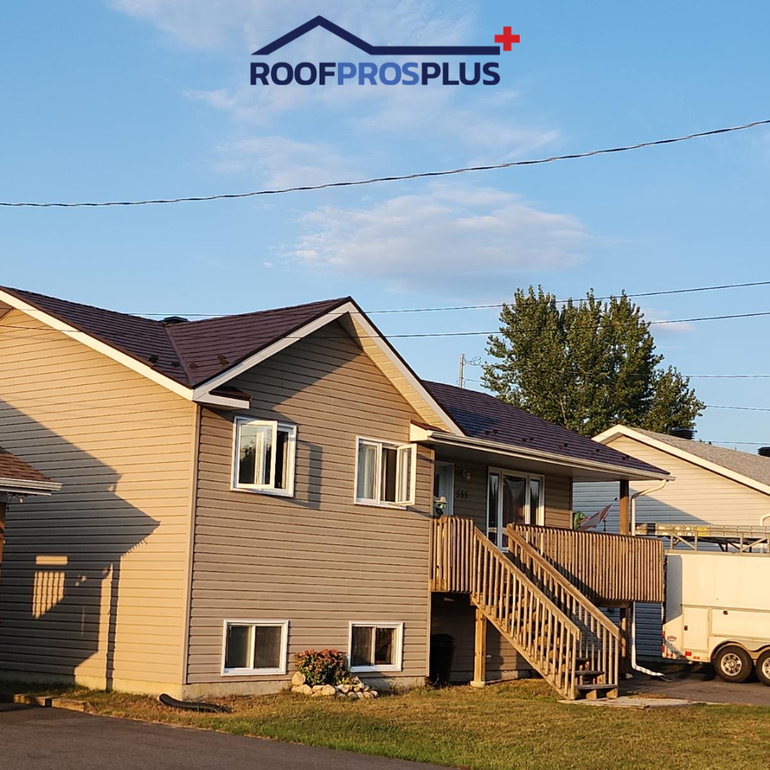 A picture of a beige two-story house with a dark metal roof, wooden porch steps, and a sunny yard. The "RoofProsPlus" logo is at the top of the photo.