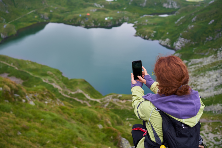 Female hiker sitting on a mountaintop taking a photo of the lake below. 