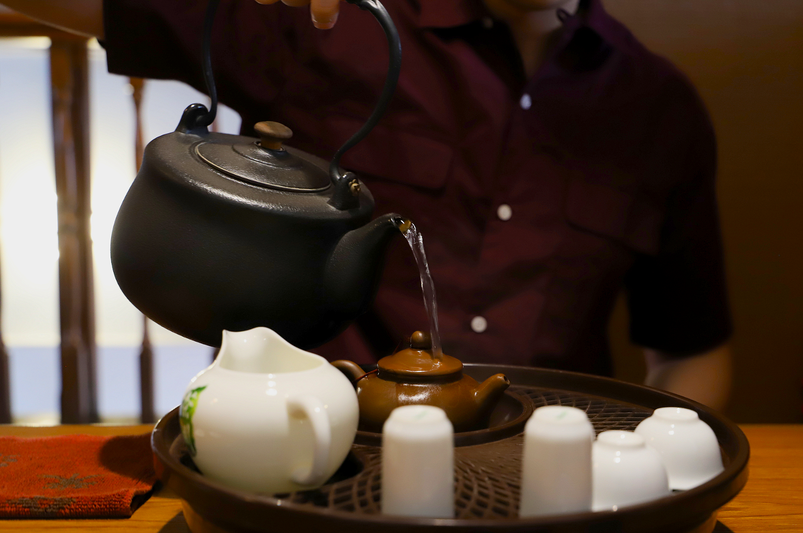 A person in a burgundy shirt pours tea from a black kettle into a small brown teapot on a tray with small white cups, creating a tranquil atmosphere.
