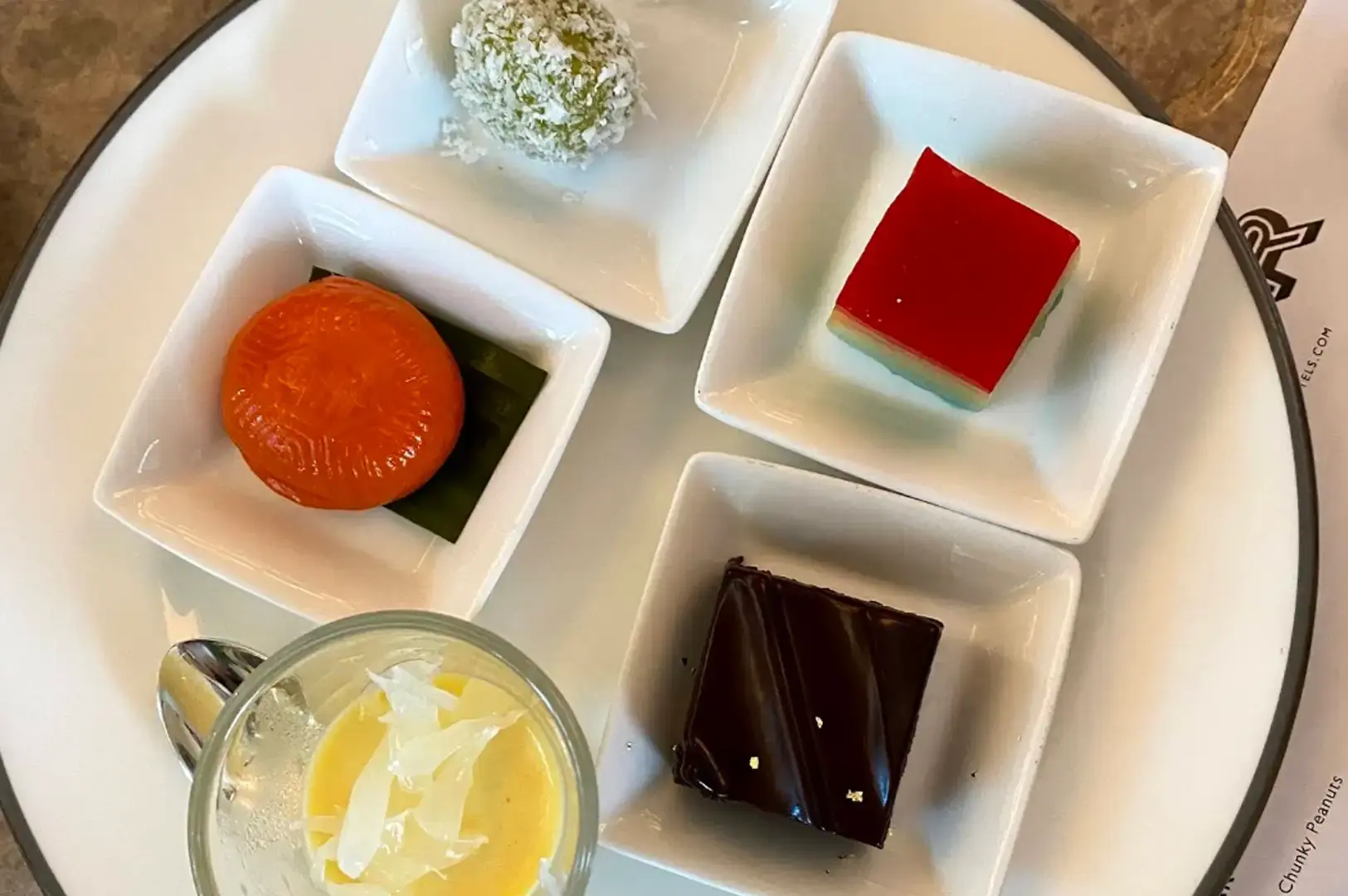 A top-down view of a round white plate holding five small square bowls containing traditional local desserts, including an orange ang ku kueh on a banana leaf, a green coconut ondeh-ondeh ball, a layered rainbow kueh lapis, a glossy dark chocolate cake square with gold leaf, and a small cup of mango pomelo dessert.