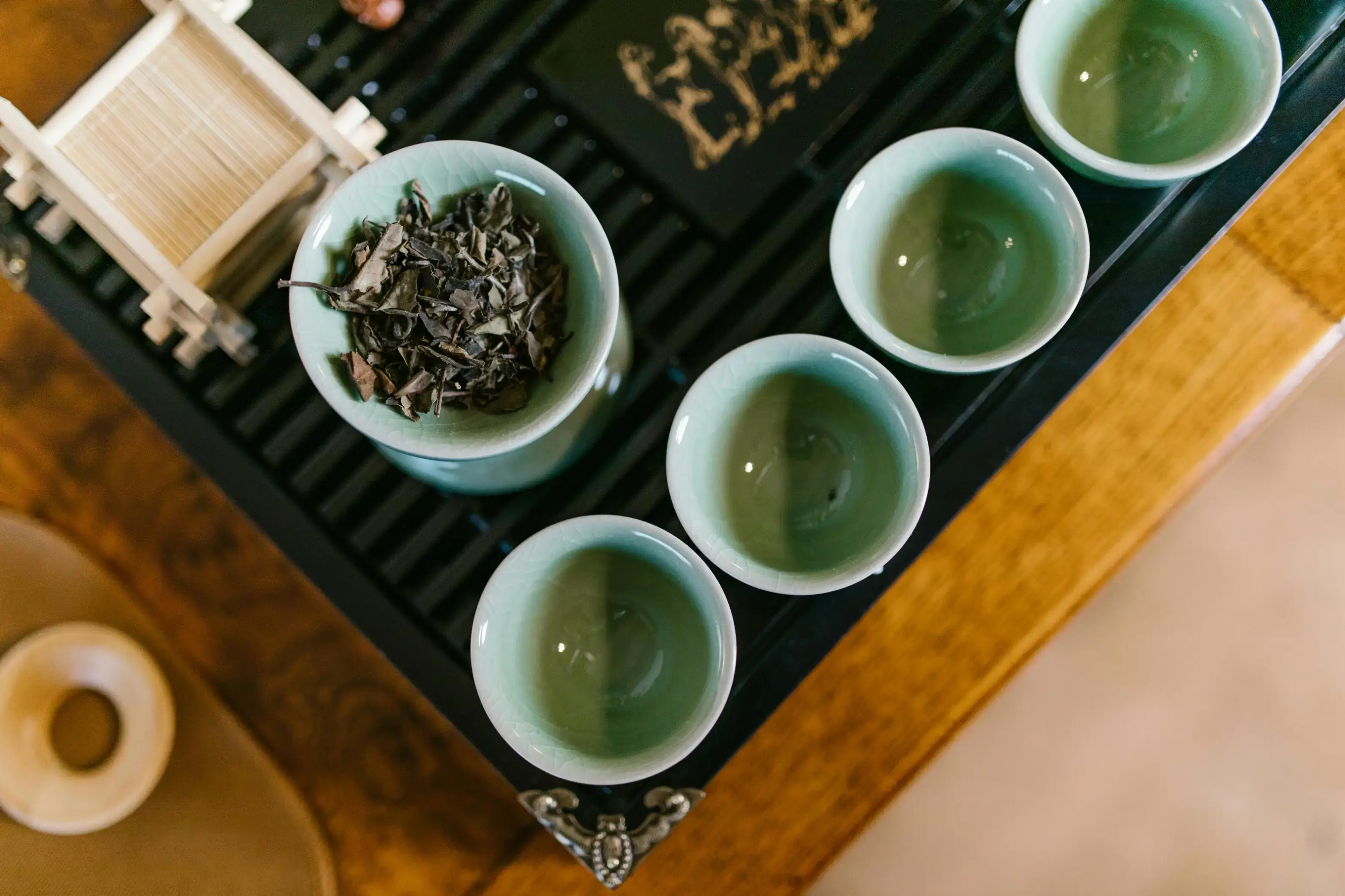 This top-down shot captures a traditional tea set with several pale green ceramic cups and a gaiwan filled with loose tea leaves. The arrangement is neatly placed on a dark, slatted tea tray atop a wooden surface, evoking a serene and ritualistic atmosphere.