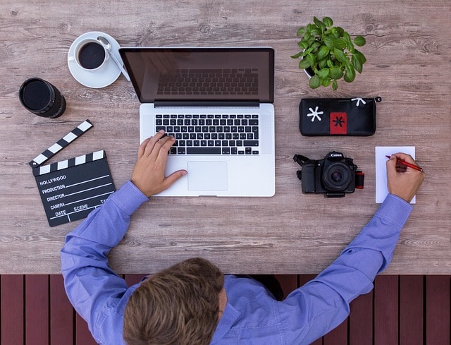 A screenwriter adapting a short story for the screen. He is writing on a notepad while typing on a laptop at his desk, surrounded by camera equipment.