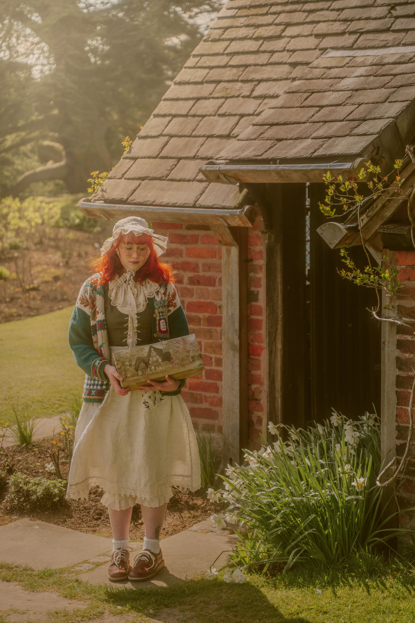 A woman in a knit sweater and pinafore dress wearing a bonnet steps out of a brick cottage