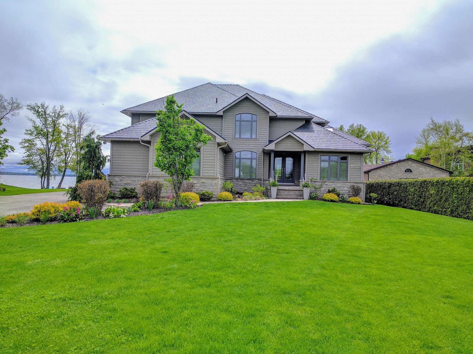 Two-story suburban house with grey siding, a metal roof and large windows, surrounded by a well-maintained lawn, shrubs, and trees, under an overcast sky.