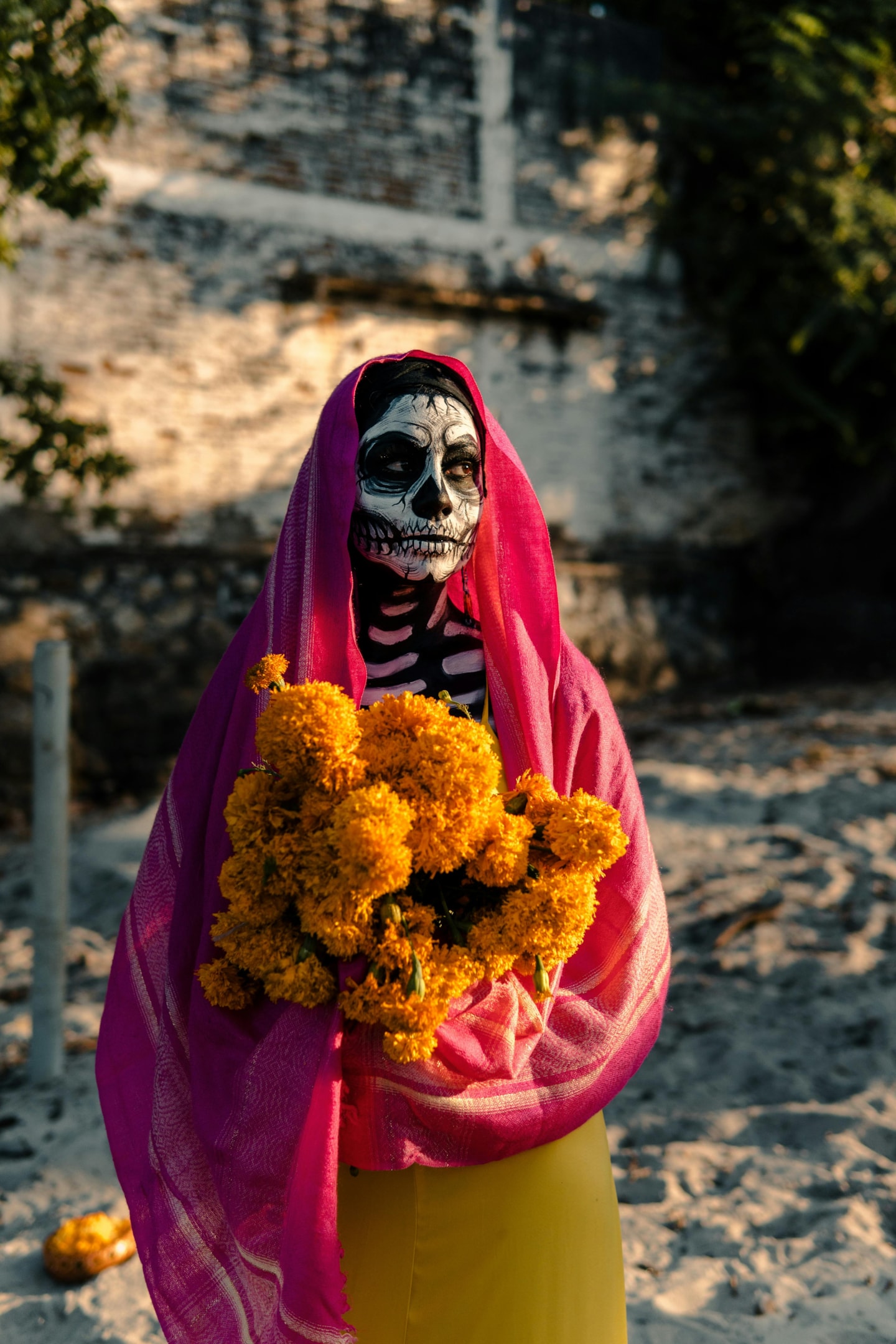A person dressed as La Catrina for Día de los Muertos, holding marigolds in a cemetery. 