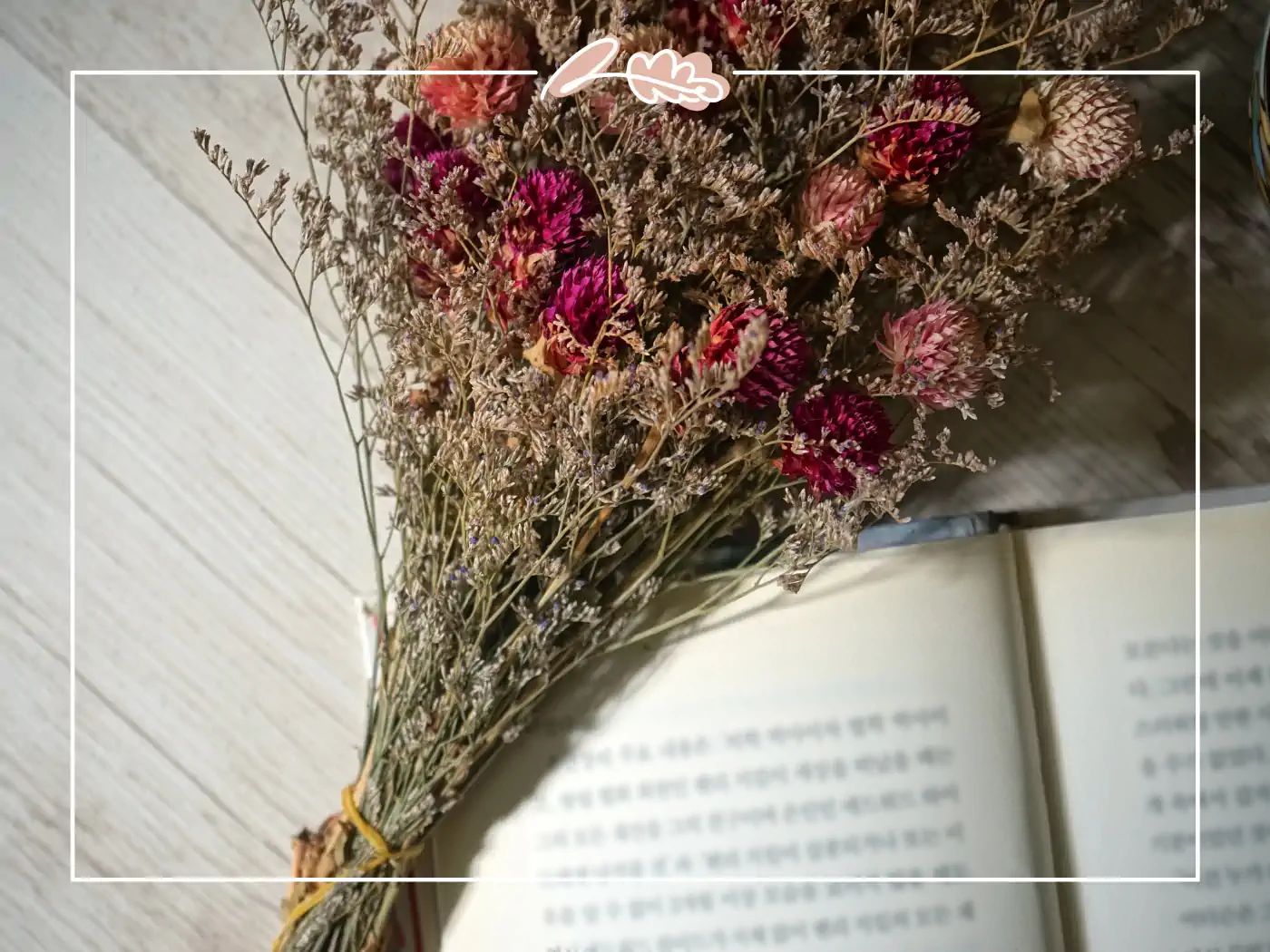 A delicate bouquet of dried flowers, featuring deep pink and light purple blooms, laid beside an open book. Fabulous Flowers and Gifts.