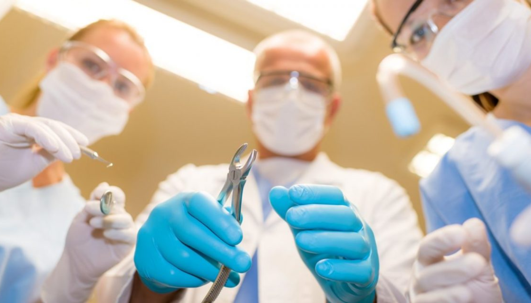 Tannlegeteam med munnbind og hansker forbereder seg på tannbehandling med instrumenter under sterilt lys, sett fra pasientens perspektiv.