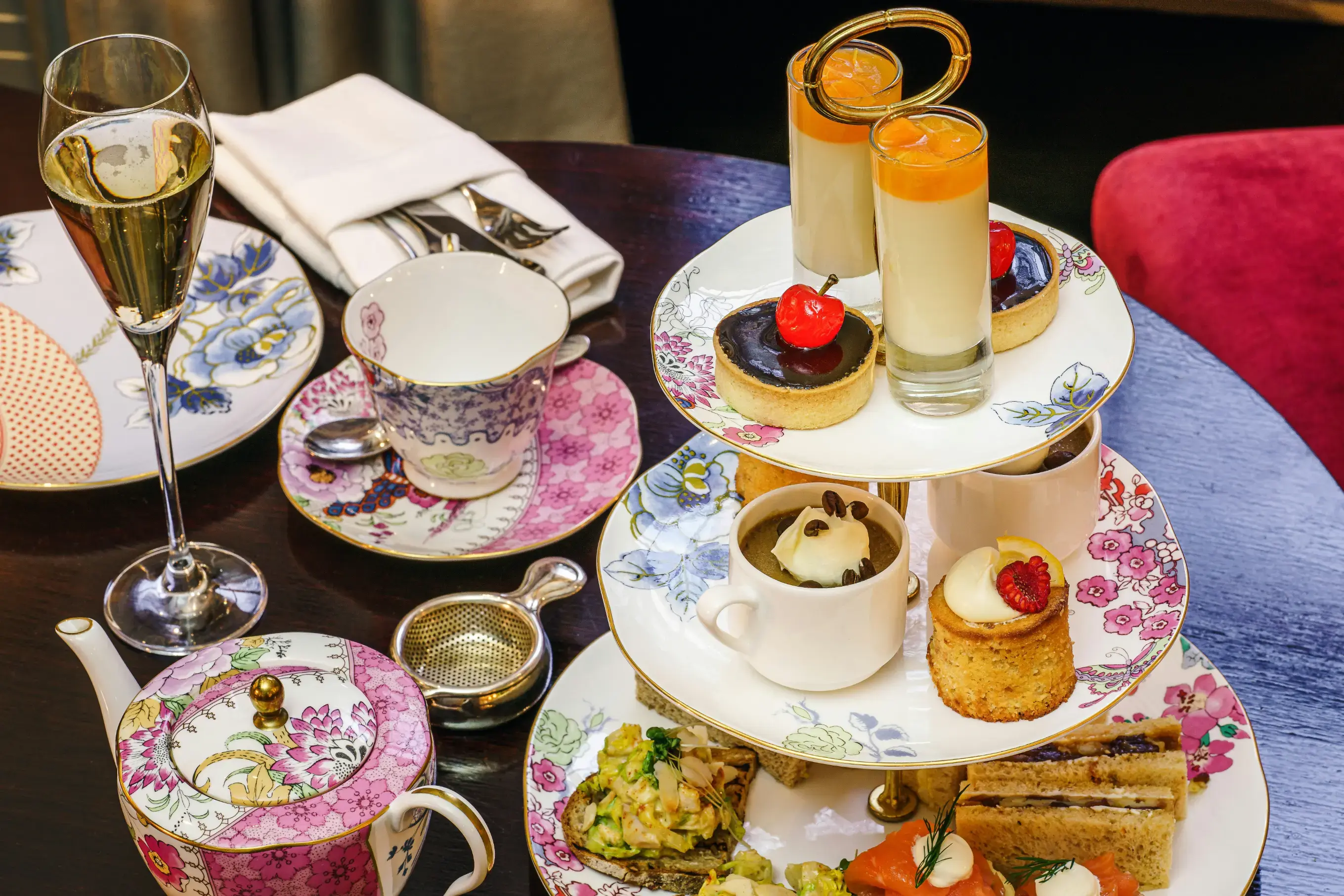 A three-tiered cake stand holds an assortment of afternoon tea treats, including finger sandwiches on the bottom tier, small cakes and parfaits in the middle, and tarts and layered desserts on top. The wooden table is also set with a floral teapot and matching teacup, a glass of champagne, and folded napkins with cutlery.