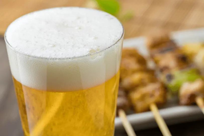 A close-up macro shot of a crisp, cold glass of draft beer with a thick, frothy head, with grilled yakitori skewers softly blurred in the background.