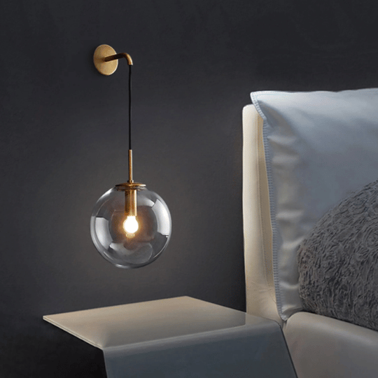 Modern wall-mounted bedside lamp with a round transparent glass globe and exposed bulb, suspended by a black cord and brass fixture, next to a bed with white bedding and a gray textured pillow.