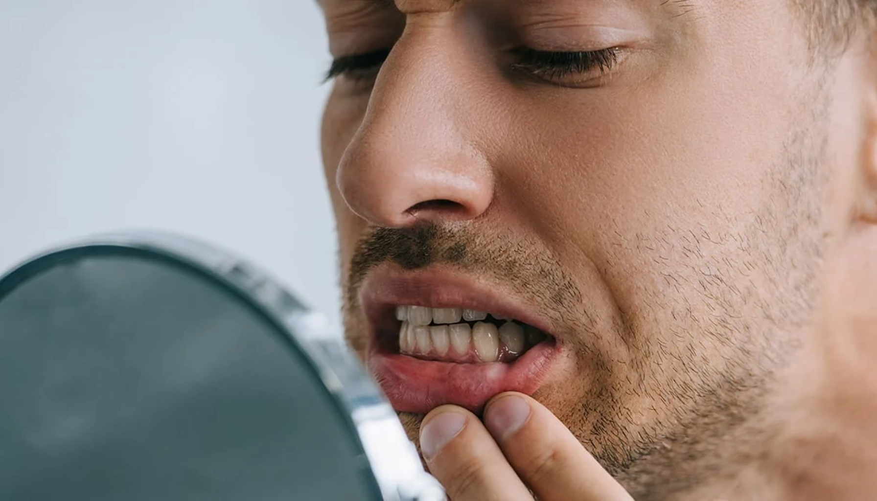 Mann ser bekymret på tennene sine i speil og undersøker underleppen med fingrene for mulig skade eller smerte i munnen.