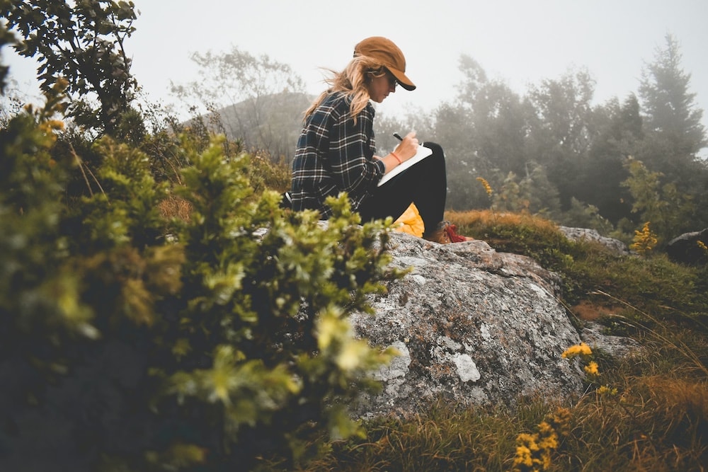 A white blond woman in a caramel-colored baseball hat, black flannel shirt with sleeves rolled up, and black pants sits on a large rock in a field amongst trees. It's a foggy day. She rests a notebook on her lap and writes in the journal as the wind blows her hair back.