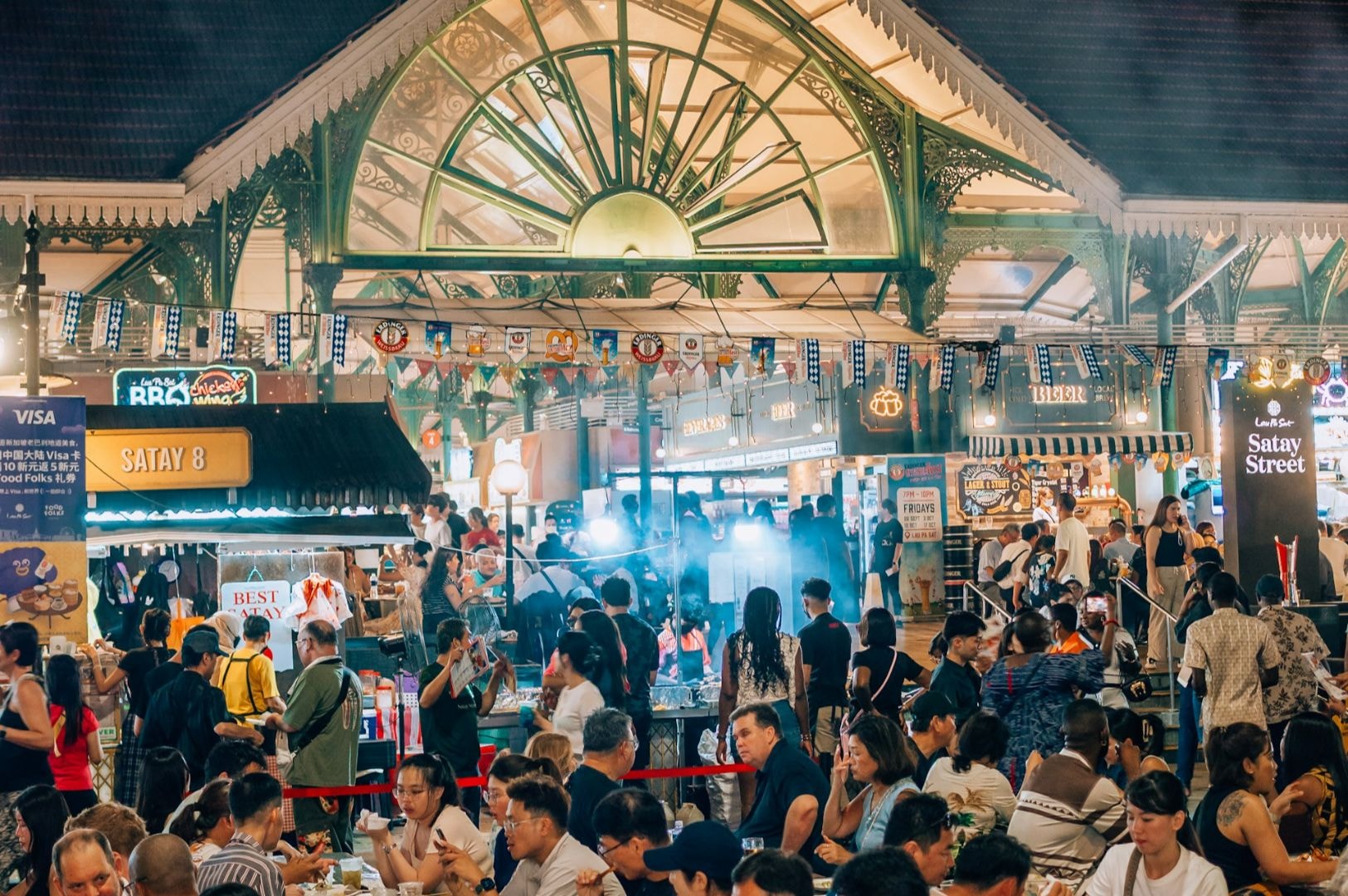 Bustling night market with diverse food stalls under a lit canopy. Crowd enjoys street food, vibrant atmosphere full of energy and social interaction.