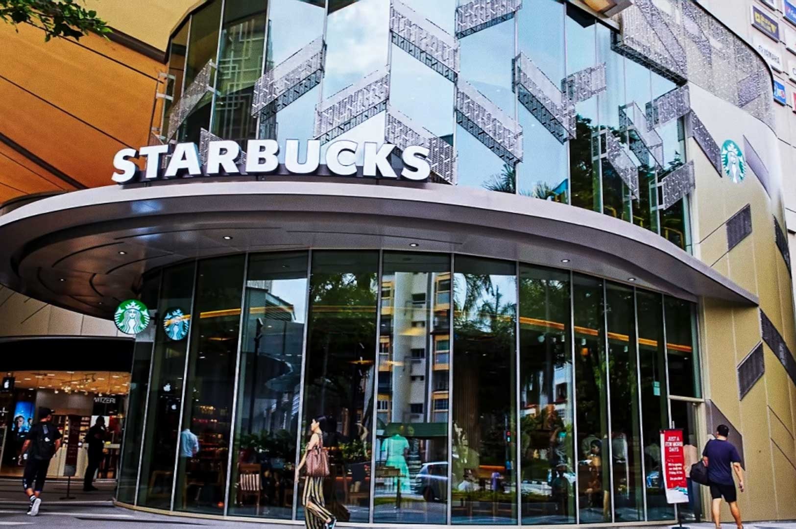 Modern Starbucks storefront at Tampines Mall with curved glass facade, bold white STARBUCKS sign above the entrance, and shoppers passing by on a busy sidewalk.