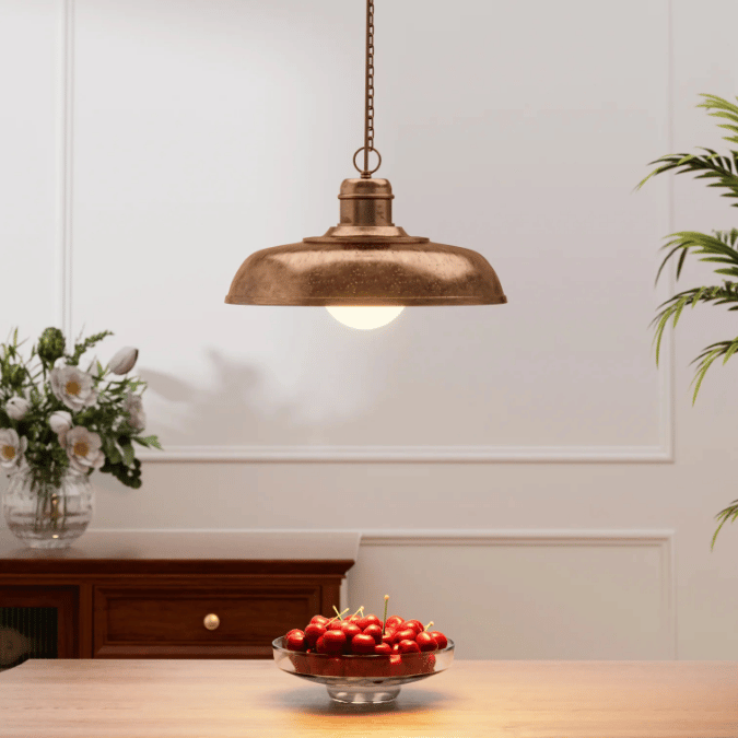 A single vintage-style copper pendant light hanging above a dining table, casting light over a bowl of cherries, with a vase of flowers and a wooden cabinet in the background.