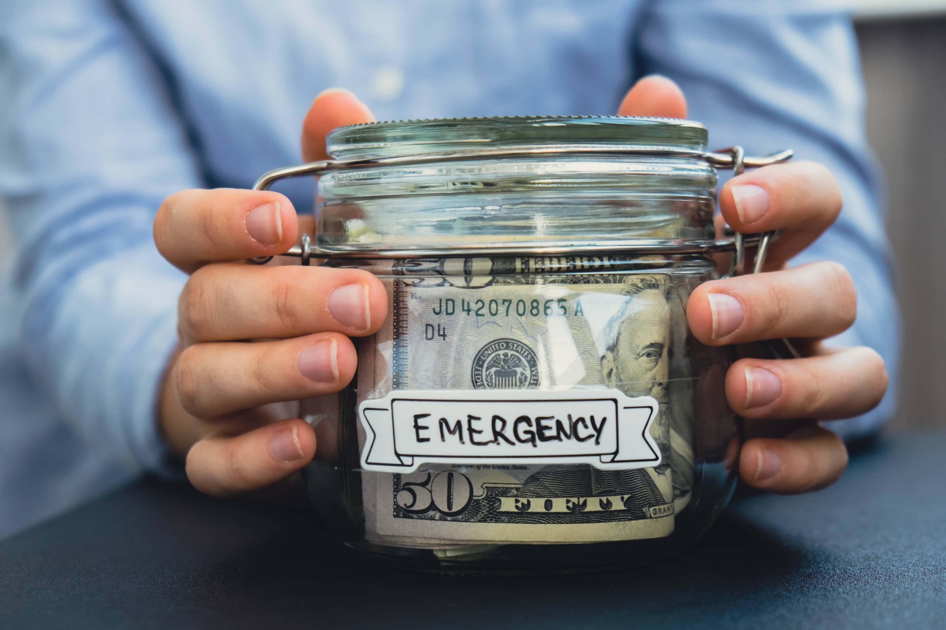 A person holding a glass jar labeled "Emergency" filled with cash, representing the importance of building an emergency fund as part of a sound financial plan.