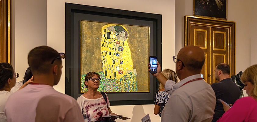 An art gallery tour stops to admire and take pictures of The Kiss, a famous Art Nouveau piece by Gustav Klimt.