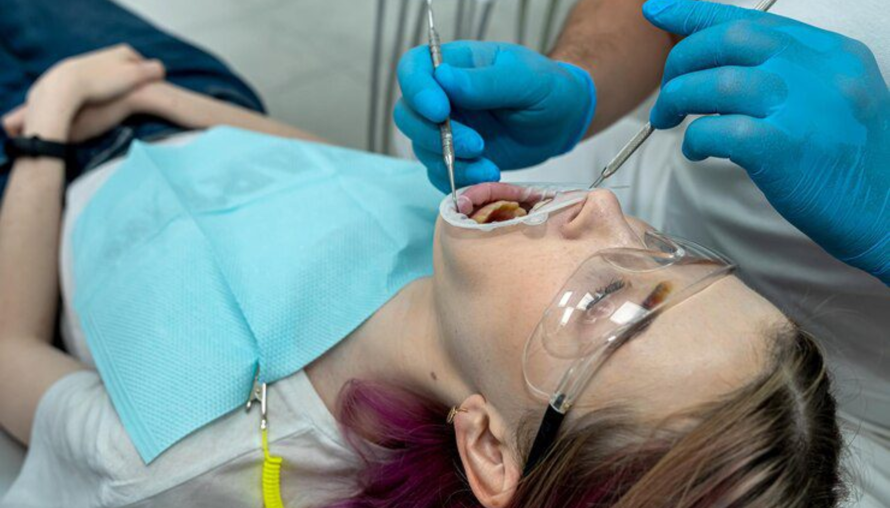 Vondt i Tann Etter Boring? Det Skal Ikke Være Slik 14 Tannlege med blå hansker undersøker munn og tenner på kvinne med beskyttelsesbriller og blå tannlegeforkle.