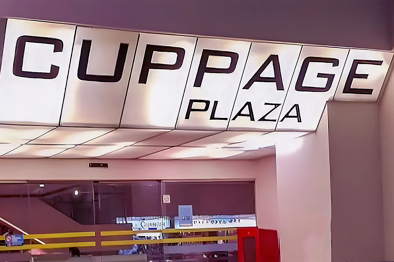 Illuminated sign reads "Cuppage Plaza" in bold letters on a ceiling panel. Below is a glass façade with a modern, commercial atmosphere.