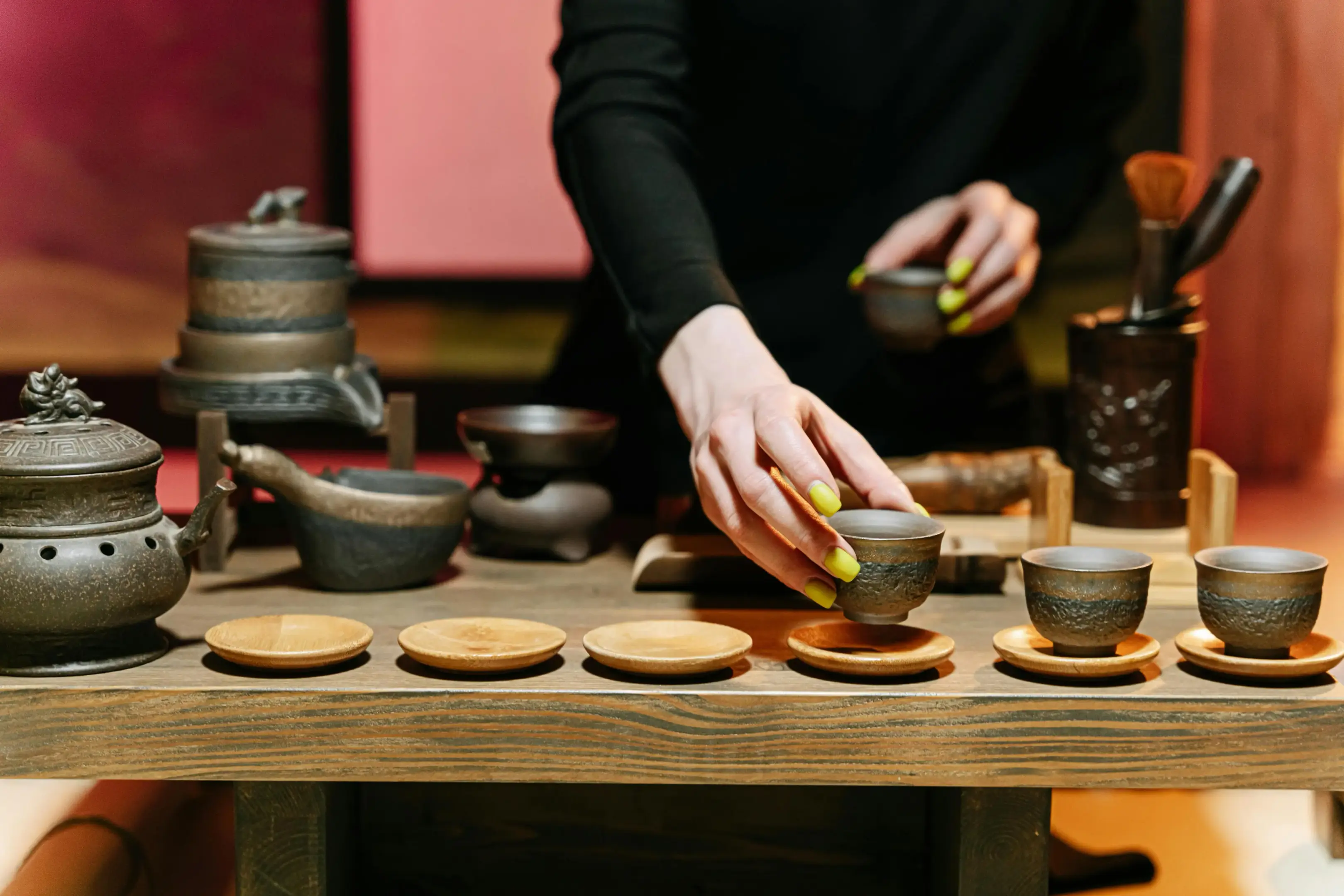 A person with bright lime-green nails carefully arranges small, rustic ceramic tea cups onto wooden saucers. The wooden table is set with an intricate collection of traditional tea-making tools, including a textured teapot and a decorative stone mill.
