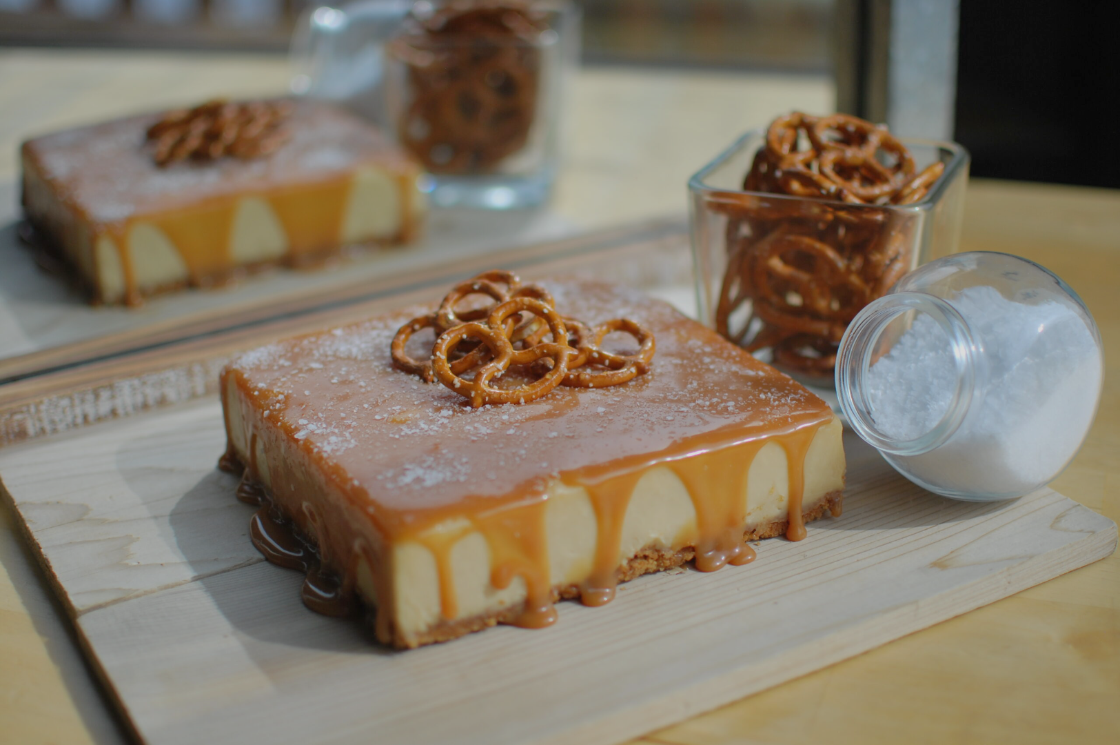 A caramel-drizzled cheesecake topped with pretzels sits on a wooden board, accompanied by a glass jar of pretzels and a spilled jar of salt.