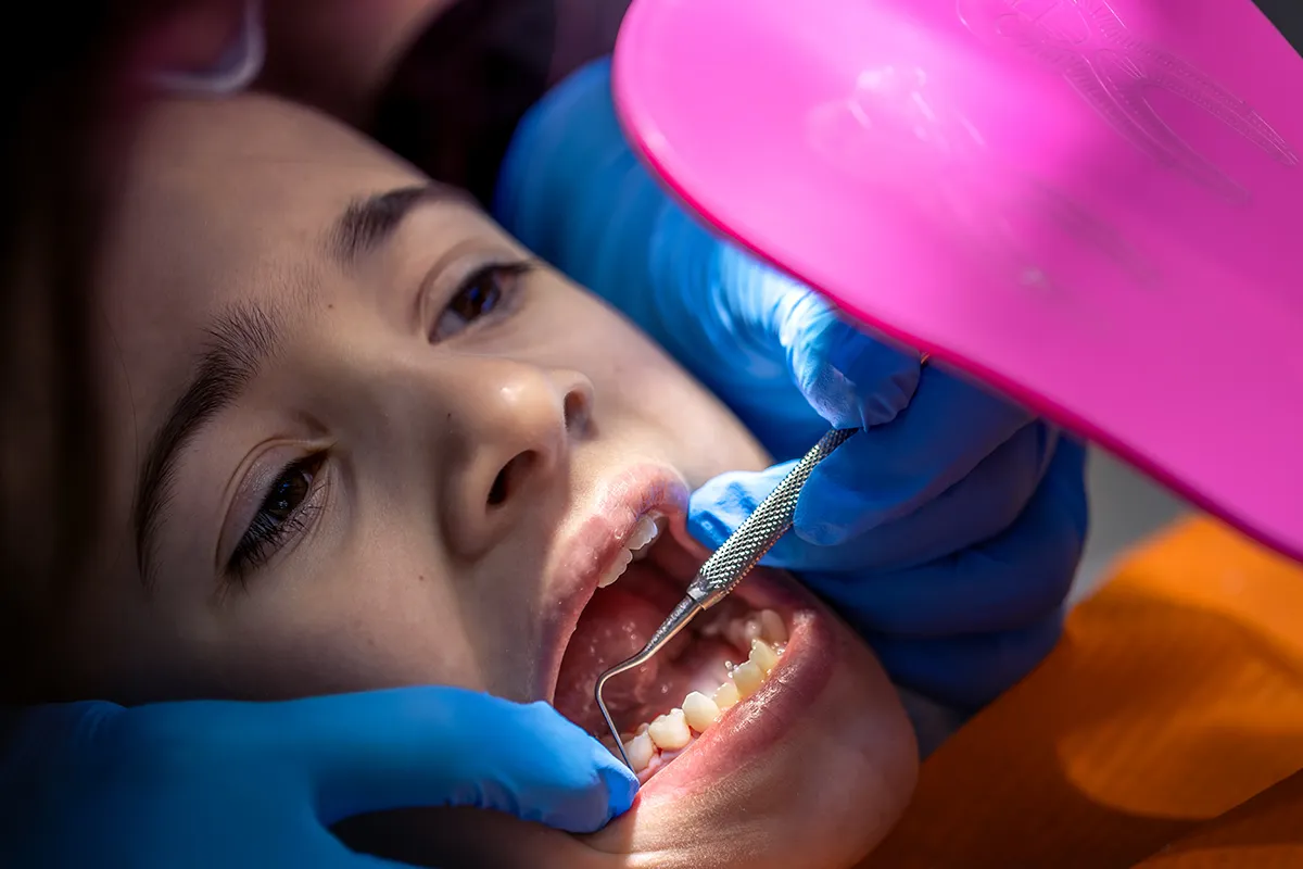 Child at dentist receiving dental examination with dental tool and pink protective shield, wearing blue dental bib.